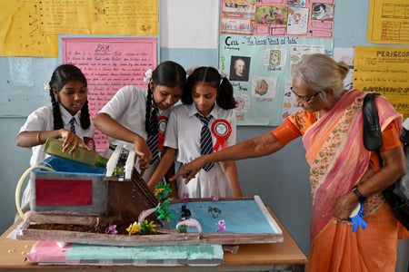 3 girls in school uniform performing a science experiment and a female visitor in orange sari visiting a classroom