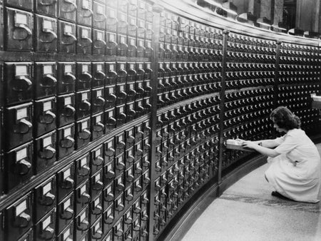 Black and white photo of a woman examining a small drawer she's pulled out, amidst rows of tiny drawers