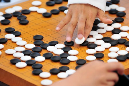 Yellow GO board with black and white pieces and two hands playing.