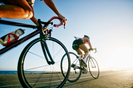 Two women bicycling in shorts and helmet on a road - only yhe wheel is showing on the left - full cyclist on right.