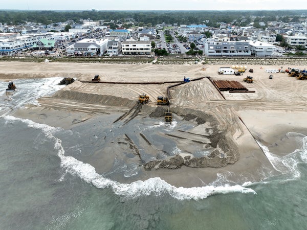 Aerial view of coastal city in background with sand being dropped and distributed in front of the beach due to erosion.