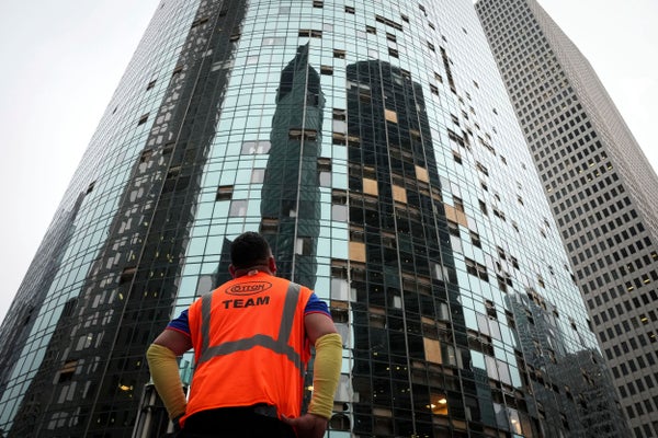 A man in orange safety jacket looks at a highrise with broken many broken windows.